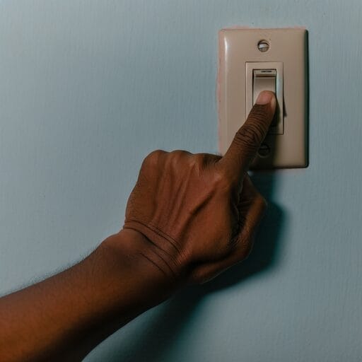 Hand reaching towards a light switch on a wall, switch in the off position, details of the switch and wall visible.
